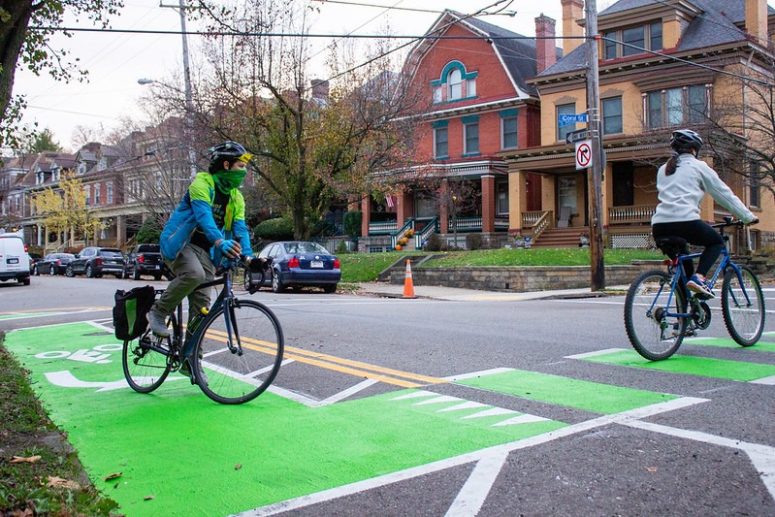 Two people on bikes cross on a painted intersection in a residential neighborhood.