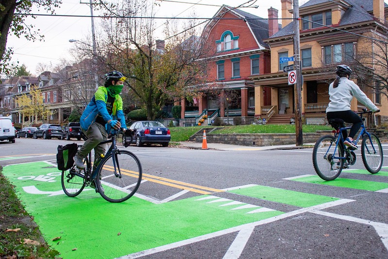 Two people on bikes cross on a painted intersection in a residential neighborhood.